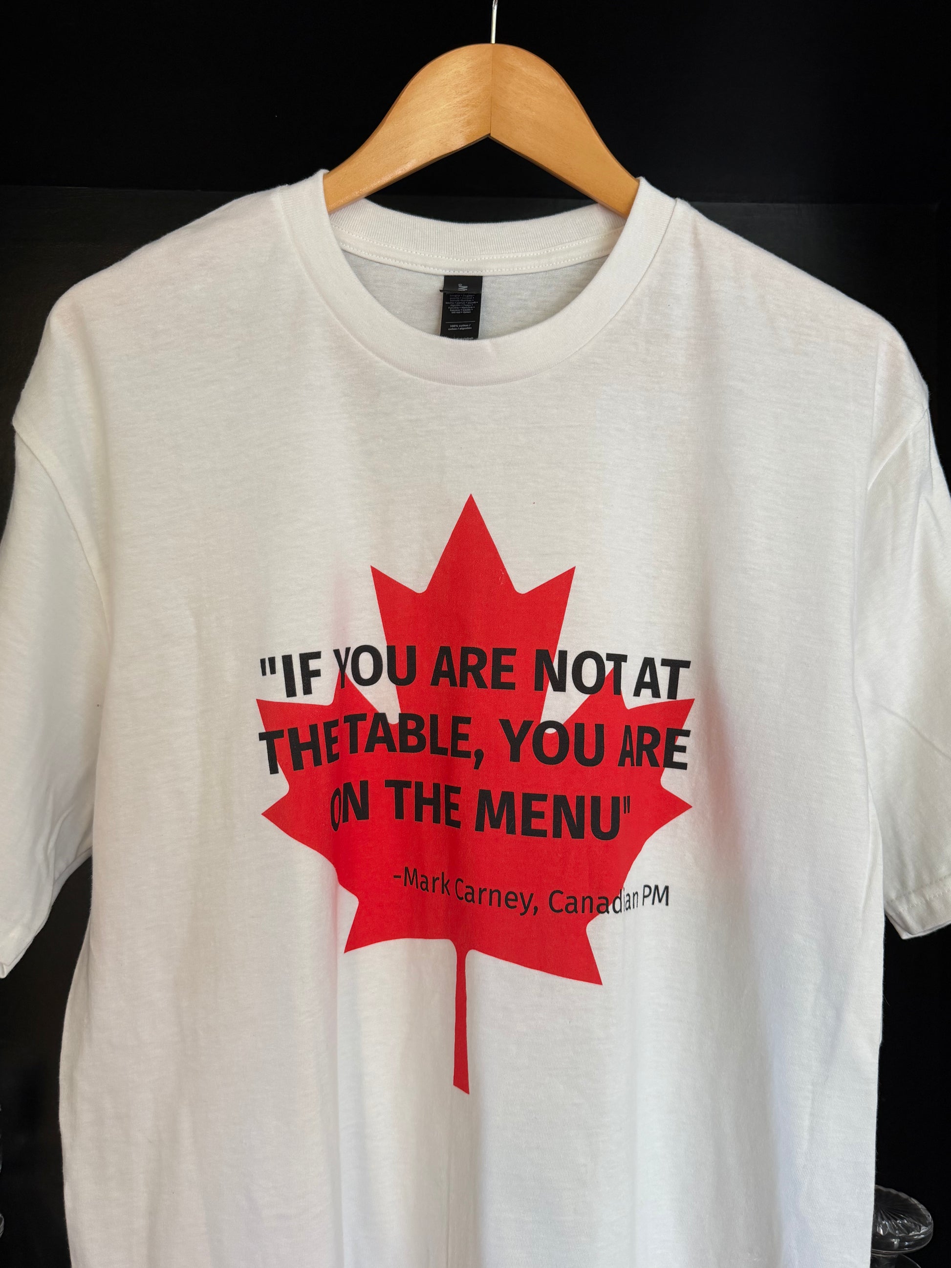 White t-shirt with lred maple leaf, overlaid in black text by the quote "If you are not at the table, you are on the menu' - attributed to Canadian PM Mark Carney