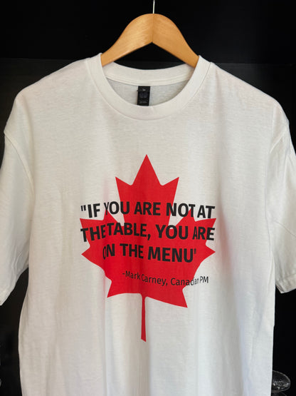 White t-shirt with lred maple leaf, overlaid in black text by the quote "If you are not at the table, you are on the menu' - attributed to Canadian PM Mark Carney