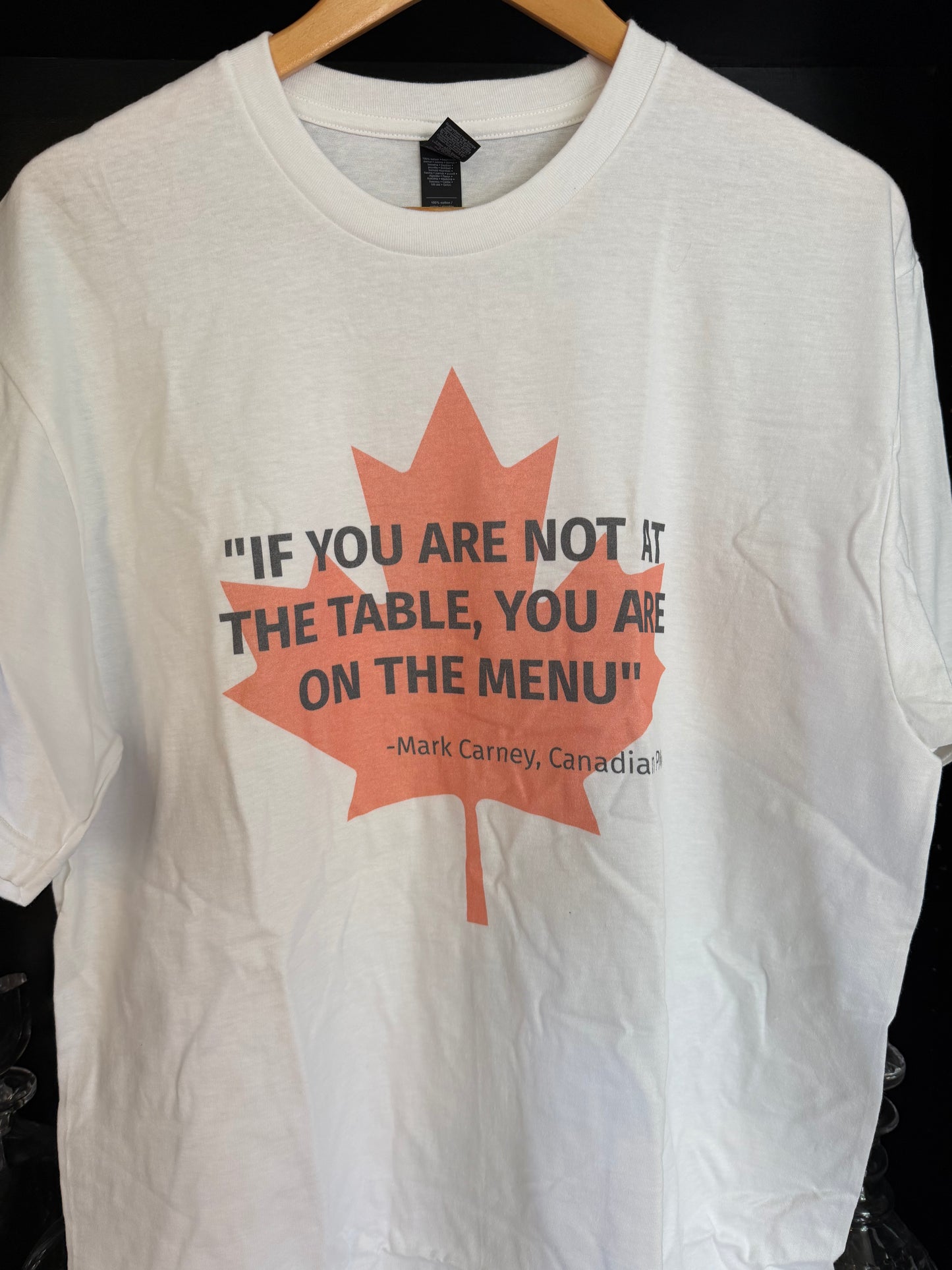 White t-shirt with light red maple leaf, overlaid in black text by the quote "If you are not at the table, you are on the menu' - attributed to Canadian PM Mark Carney