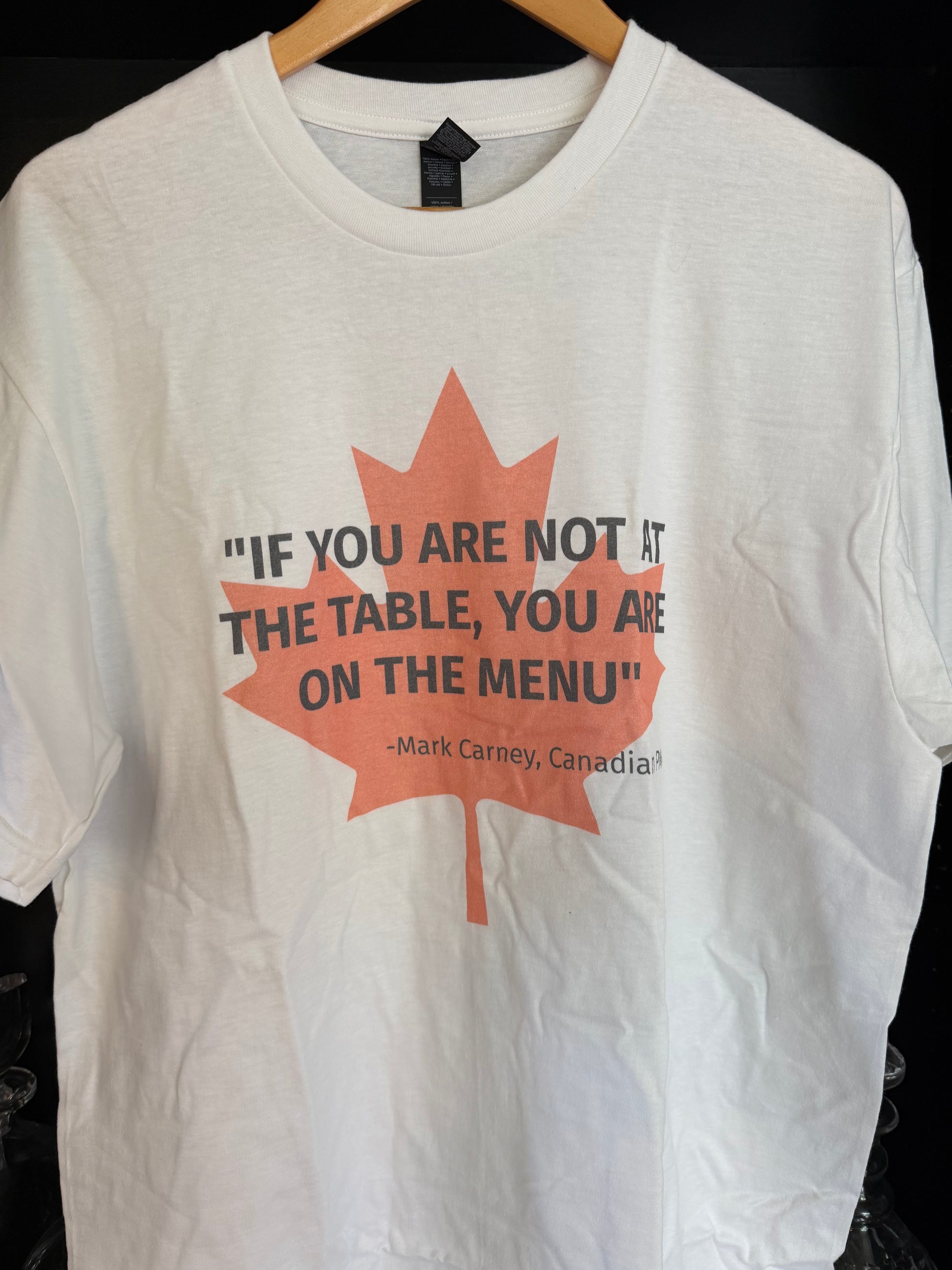 White t-shirt with light red maple leaf, overlaid in black text by the quote "If you are not at the table, you are on the menu' - attributed to Canadian PM Mark Carney
